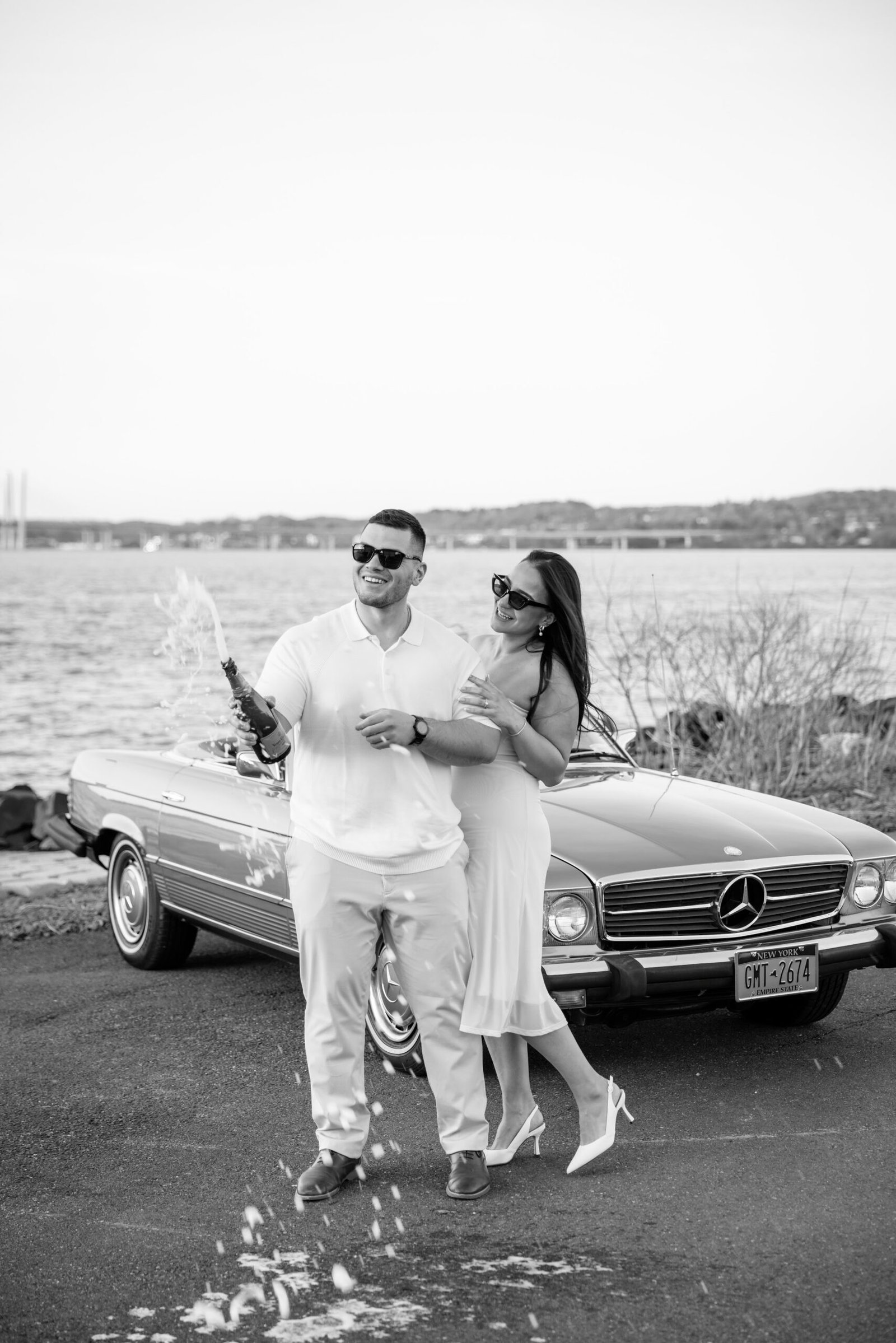 Black and white photograph of the couple celebrating with champagne beside a classic convertible